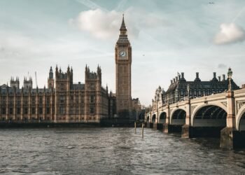 The Houses of Parliament and Elizabeth Tower clock rise beside the River Thames, with the arched Westminster Bridge on the right under a cloudy sky.
