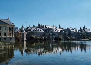 Historic brick parliament buildings with pointed towers and a classical museum reflected in a calm pond under a clear blue sky.