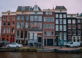 A row of tall, narrow brick canal houses with bicycles, parked cars and moored boats along a canal under a pale evening sky.