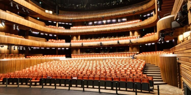View from the stage of an empty auditorium with rows of orange seats, three curved wooden balcony tiers, and stage lights hanging overhead.