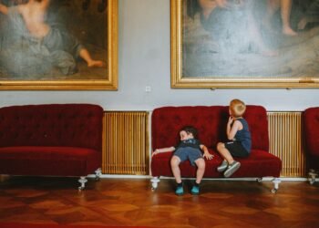Two young boys sit on a red velvet sofa in an ornate room with parquet floors, below large gilt-framed paintings, one slouching sleepily.