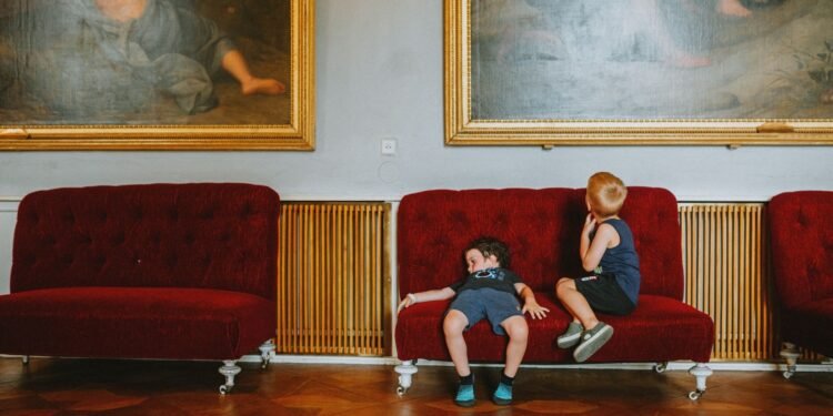 Two young boys sit on a red velvet sofa in an ornate room with parquet floors, below large gilt-framed paintings, one slouching sleepily.