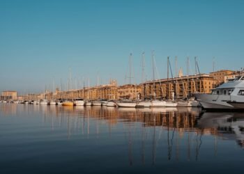 Rows of white sailboats and a large motor cruiser line a still harbour, their masts reflected in the water beneath sunlit stone buildings and clear sky.