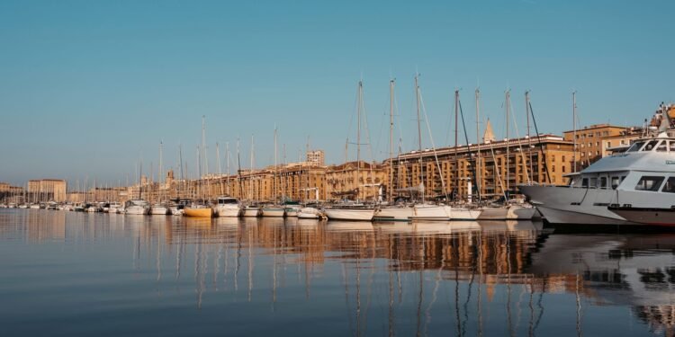 Rows of white sailboats and a large motor cruiser line a still harbour, their masts reflected in the water beneath sunlit stone buildings and clear sky.