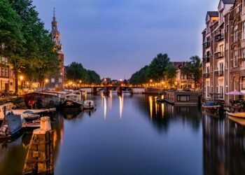 A calm canal at dusk with moored boats and houseboats, brick buildings, trees, glowing street lamps, a bridge and a historic church-like tower.