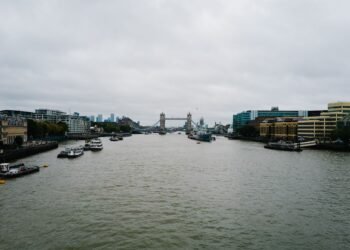 A wide river under a grey overcast sky, with moored boats and riverside buildings on both banks and Tower Bridge in the distance.
