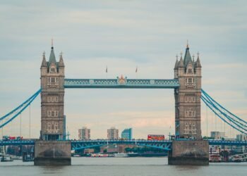 Tower Bridge's two stone towers and blue walkways span the River Thames. A red double-decker bus crosses and boats are moored below a cloudy sky.